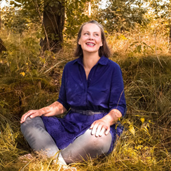Maria Ehrenberg sitzt lachend auf einem Waldboden.