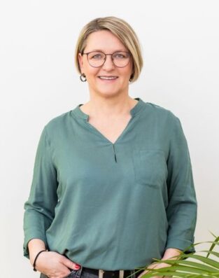 Porträt von Manuela Kind vor hellem Hintergrund. Sie trägt eine grüne Bluse, lächelt freundlich und wirkt nahbar und kompetent.